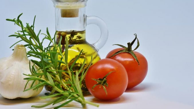 Gemüse, daneben eine Flasche hochwertiges Olivenöl, Tomaten und frische Kräuter auf einem Tisch.